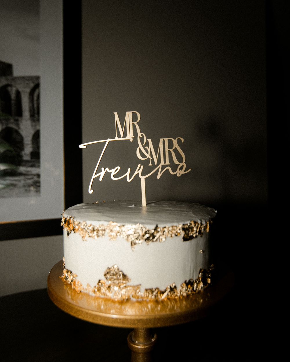 An elegant white wedding cake with gold accents and 'Mr & Mrs' topper displayed in dark lighting.