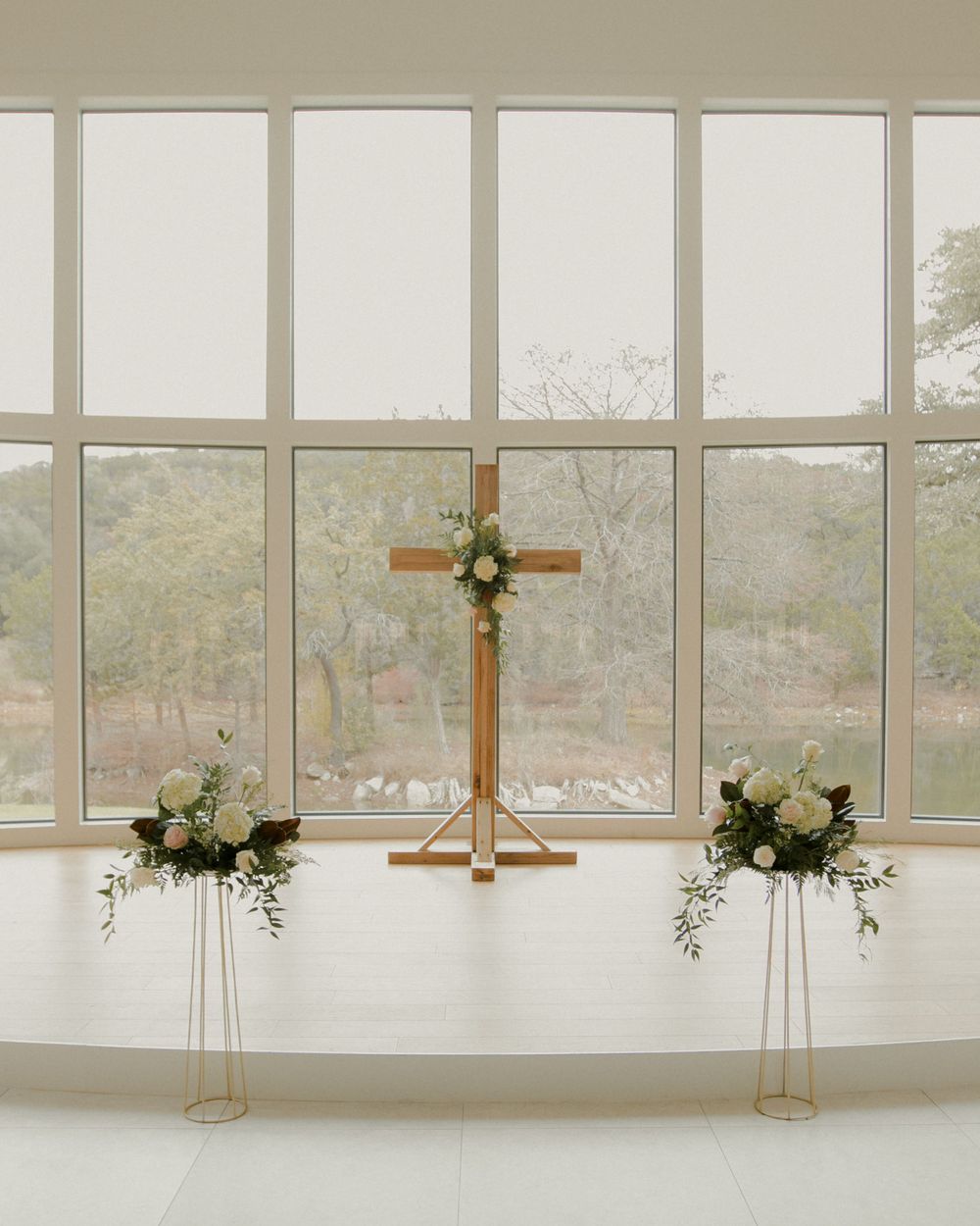 Modern white chapel interior with wooden crosses and floral arrangements against large windows.