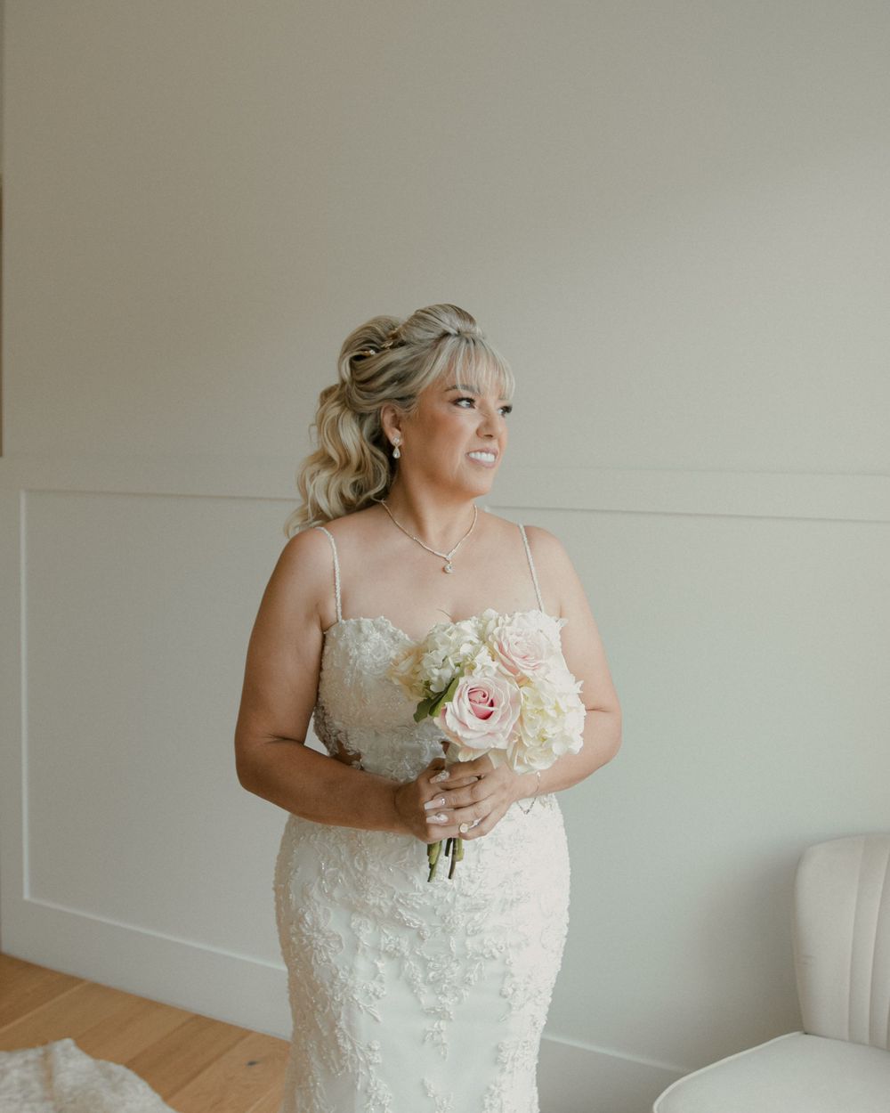 Bride in lace dress holds white bouquet while posing against light wall.
