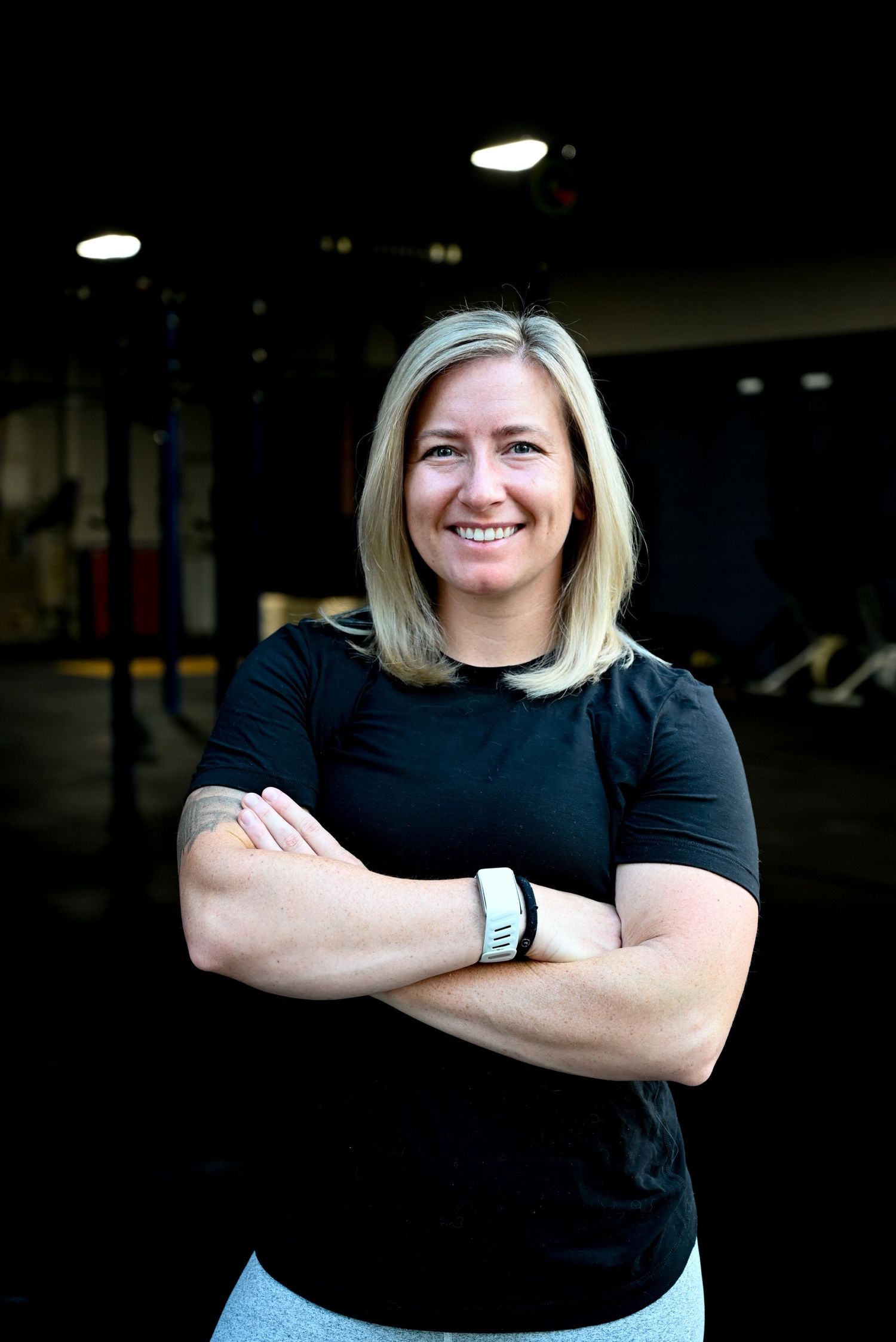 Athletic woman in black shirt standing with crossed arms in gym setting.