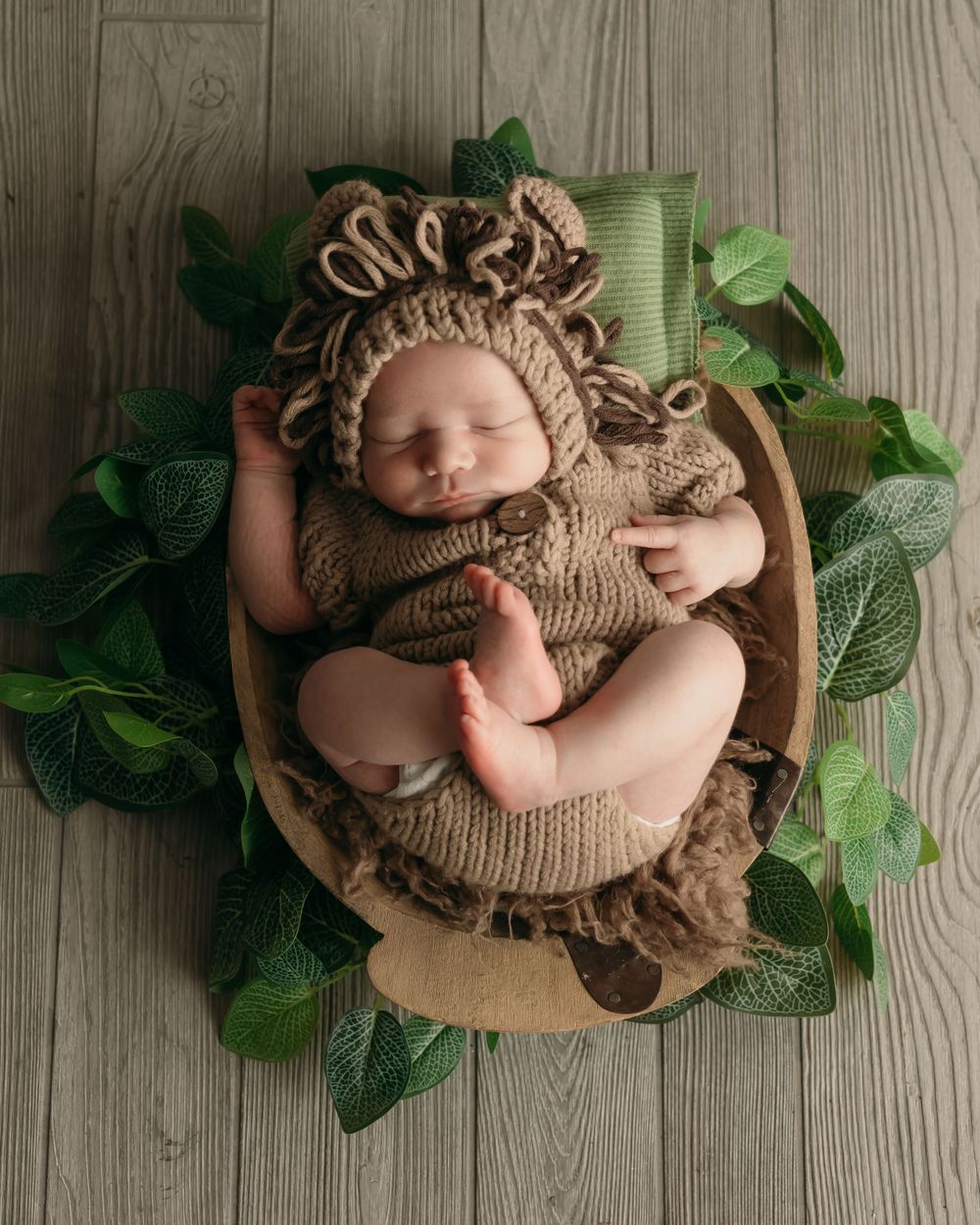 Sleeping newborn in knit bear outfit surrounded by green leaves in wooden bowl.
