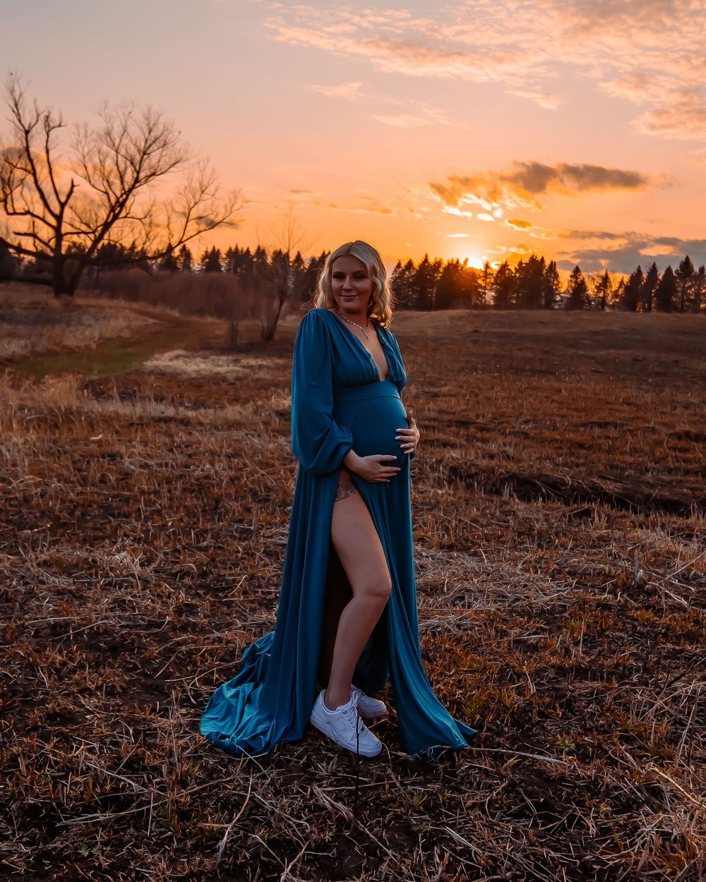 Stunning maternity pose in a flowing blue gown against a golden sunset backdrop.