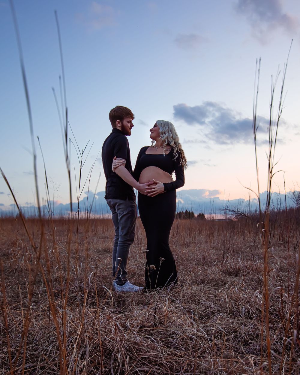 Intimate maternity portrait among tall grasses during sunset in a rural field.