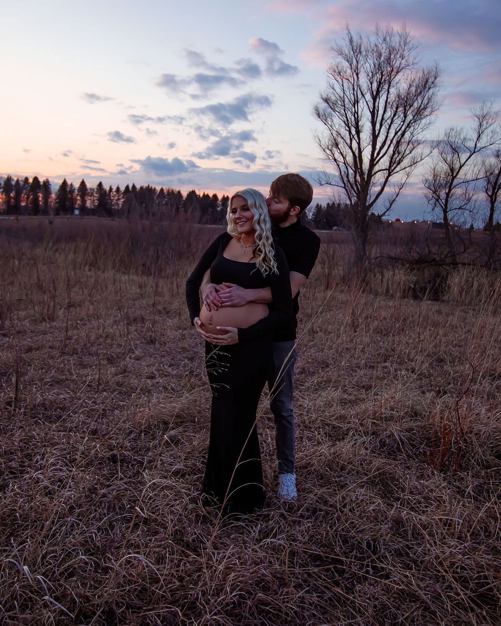 Sweet maternity moment captured in a field at dusk with moody lighting.