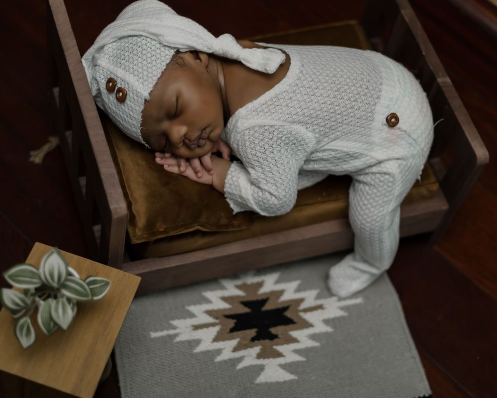 Baby sleeping peacefully in white knit outfit on brown cushion with geometric rug below.