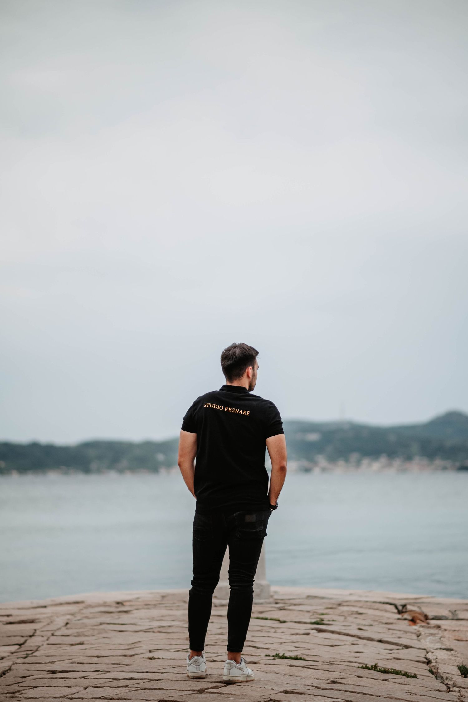 Person in black attire standing on a rocky cliff overlooking a serene waterfront landscape.