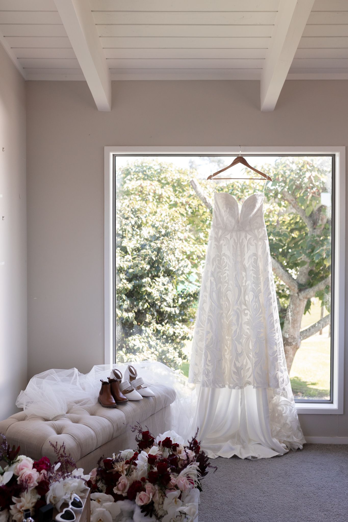 A white lace wedding dress hangs elegantly by a window with shoes and floral arrangements displayed below.