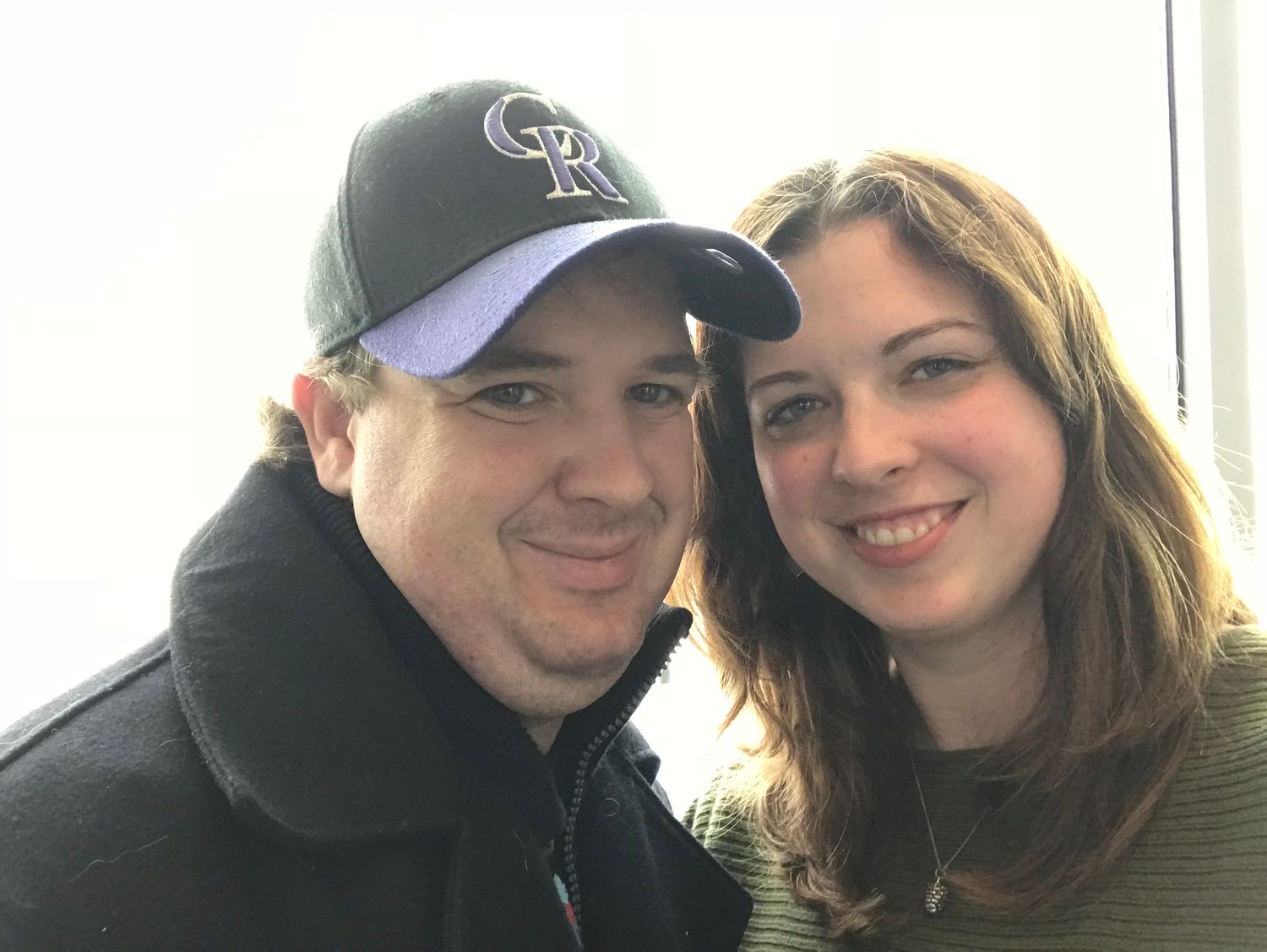 Two people in casual attire pose together for a selfie with one wearing a baseball cap.