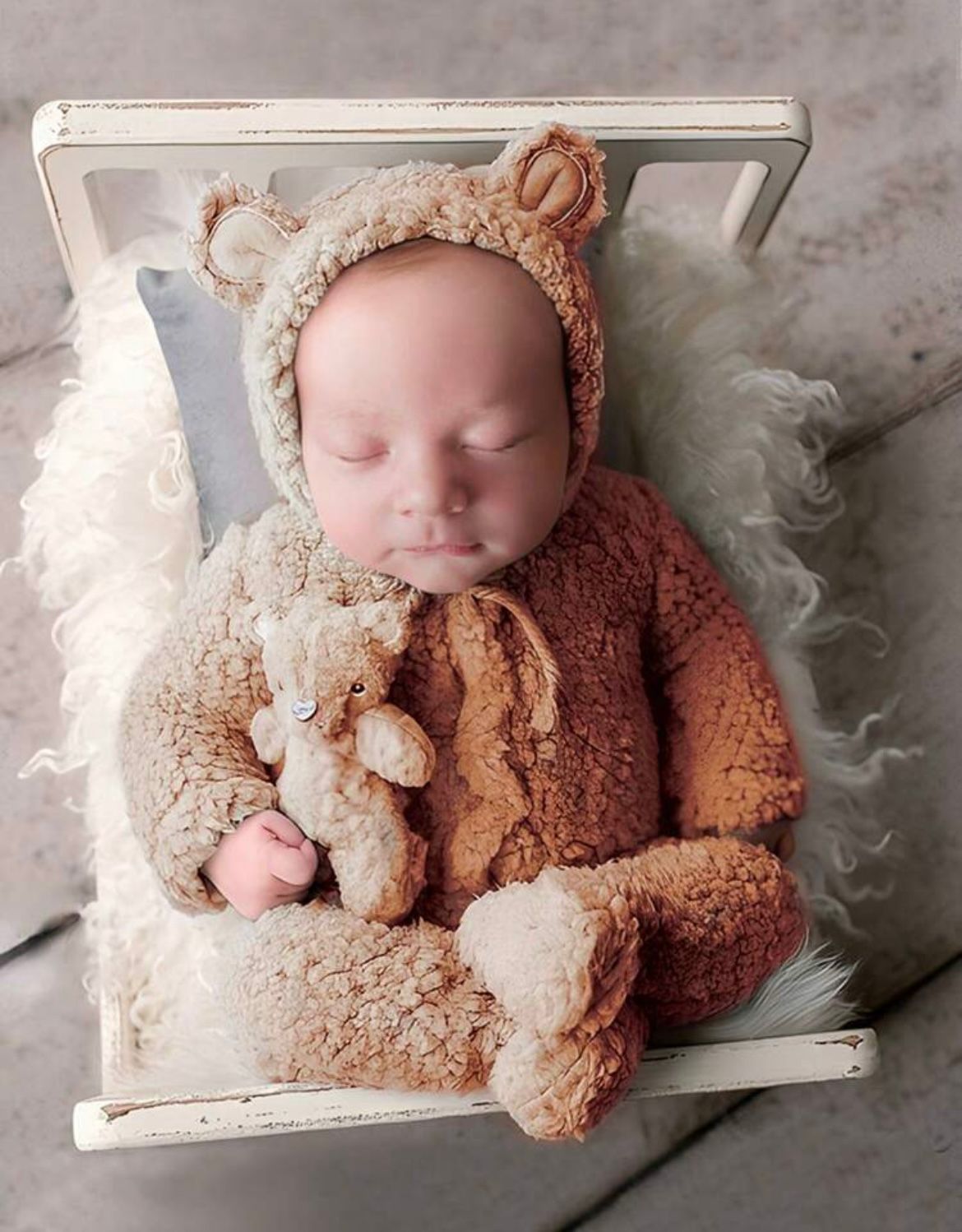 Sleeping newborn in brown teddy bear outfit lying on white vintage frame.