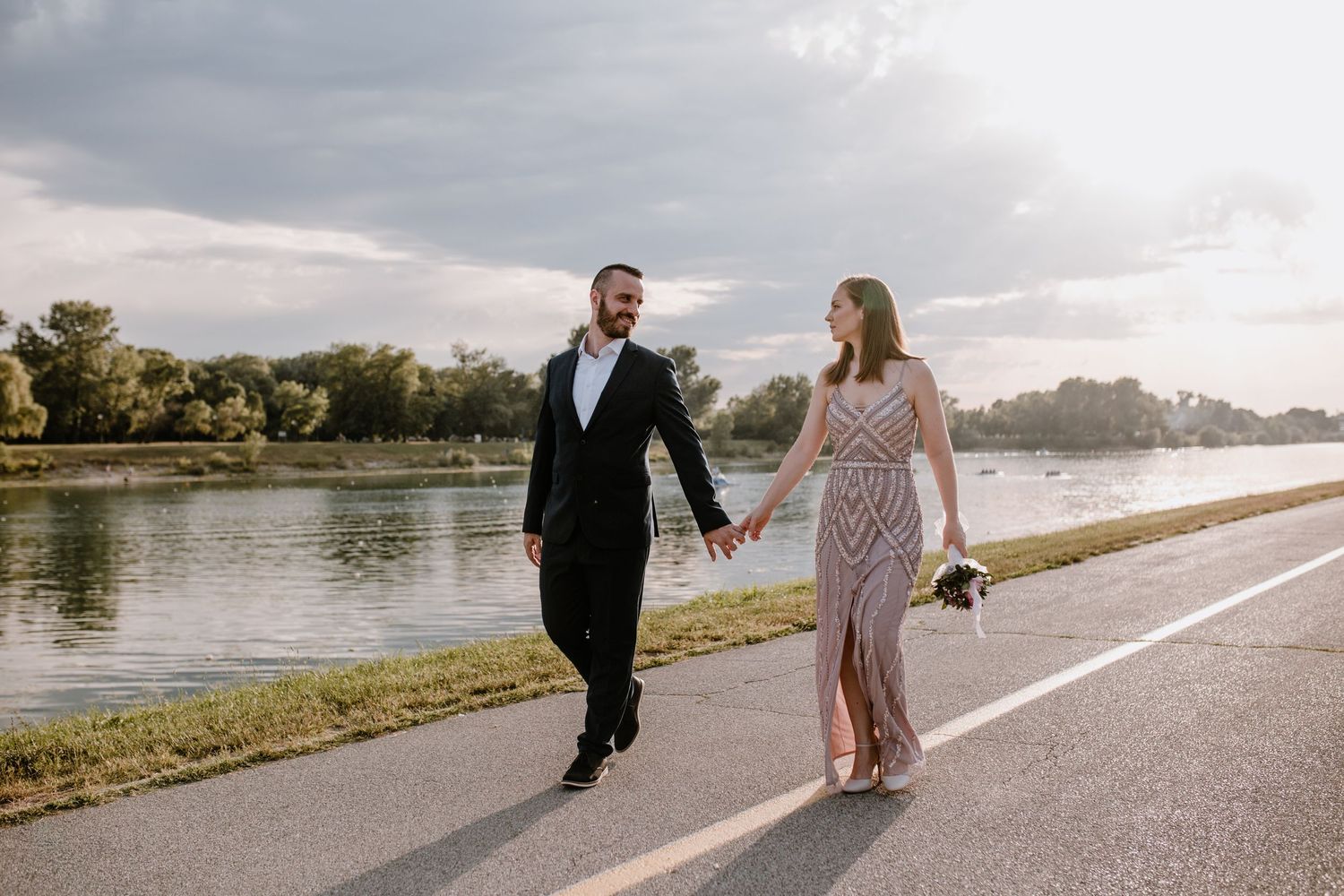 Couple walking hand in hand along riverside path during golden hour.