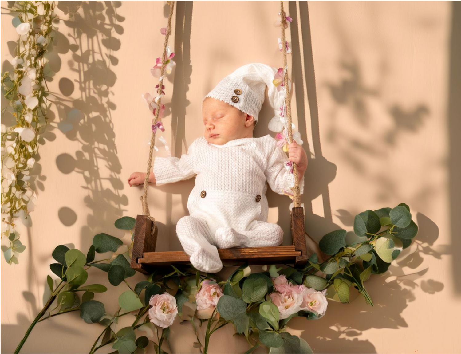 A baby wearing white winter clothing sits on a wooden swing decorated with pink roses and green vines.