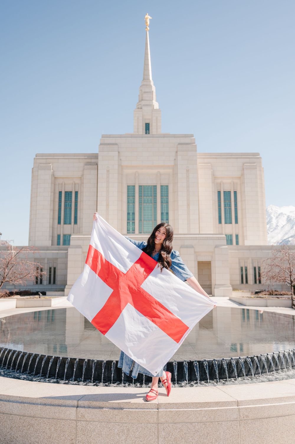 English flag being held in front of white Mormon temple building with snow-capped mountains in background.