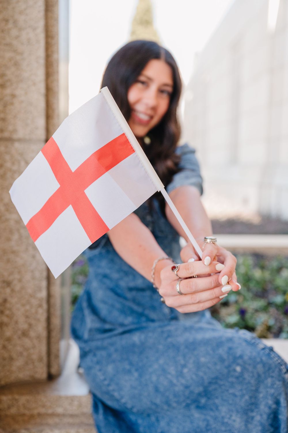A St George's Cross flag of England being waved while sitting on stone steps with greenery in the background.