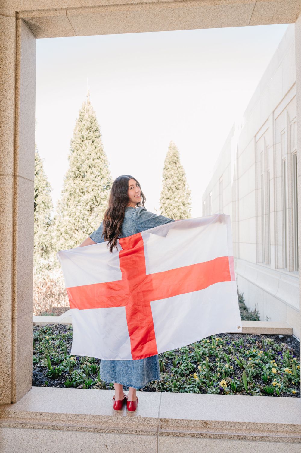 Someone draped in an England flag stands in a stone archway wearing denim and red shoes against white walls and cypress trees.