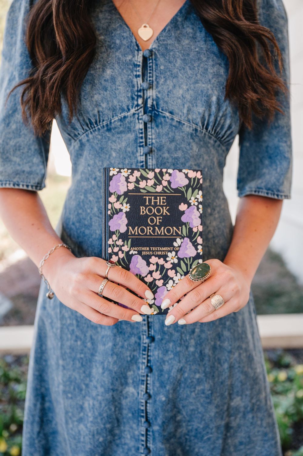 A person in a denim dress holds a floral-covered Book of Mormon against their body.