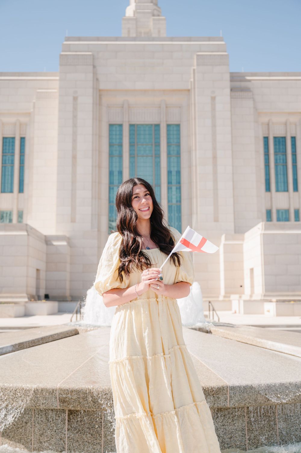 Figure in light yellow dress stands with Canadian flag in front of white stone fountain and temple building on sunny day.