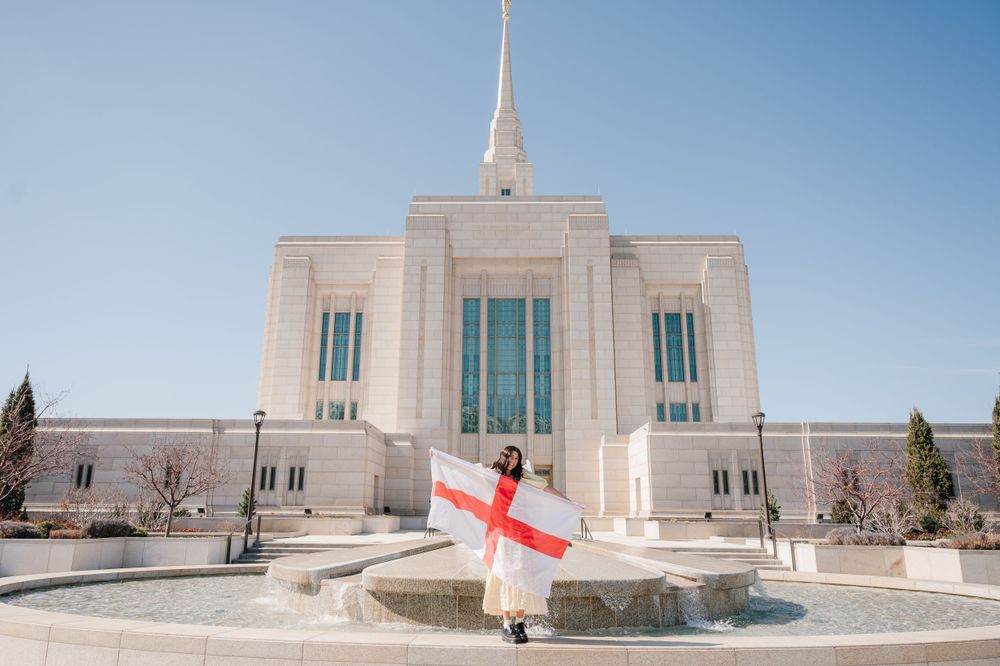 A white Mormon temple with an England flag displayed in front of the entrance on a sunny day.