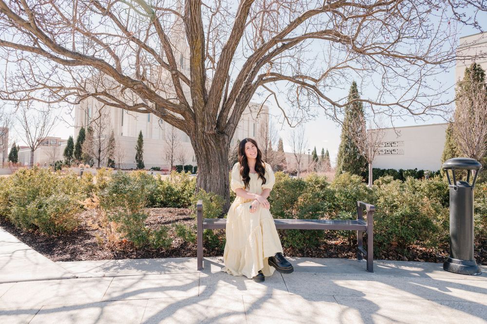 A person in a flowing cream dress stands beneath a bare winter tree in a manicured garden setting.