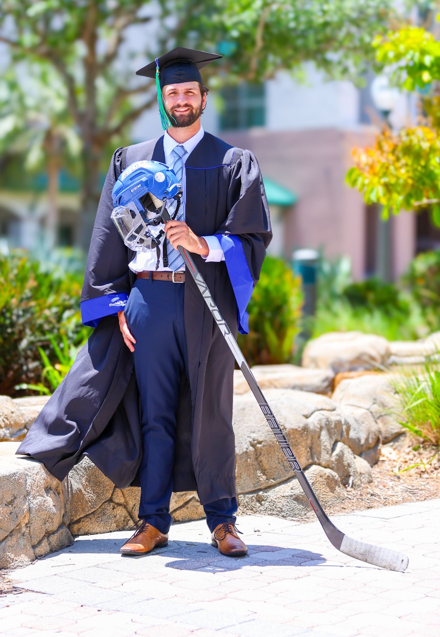 Graduate in cap and gown poses with hockey stick outdoors on sunny day. Florida Gulf Coast University, Florida
