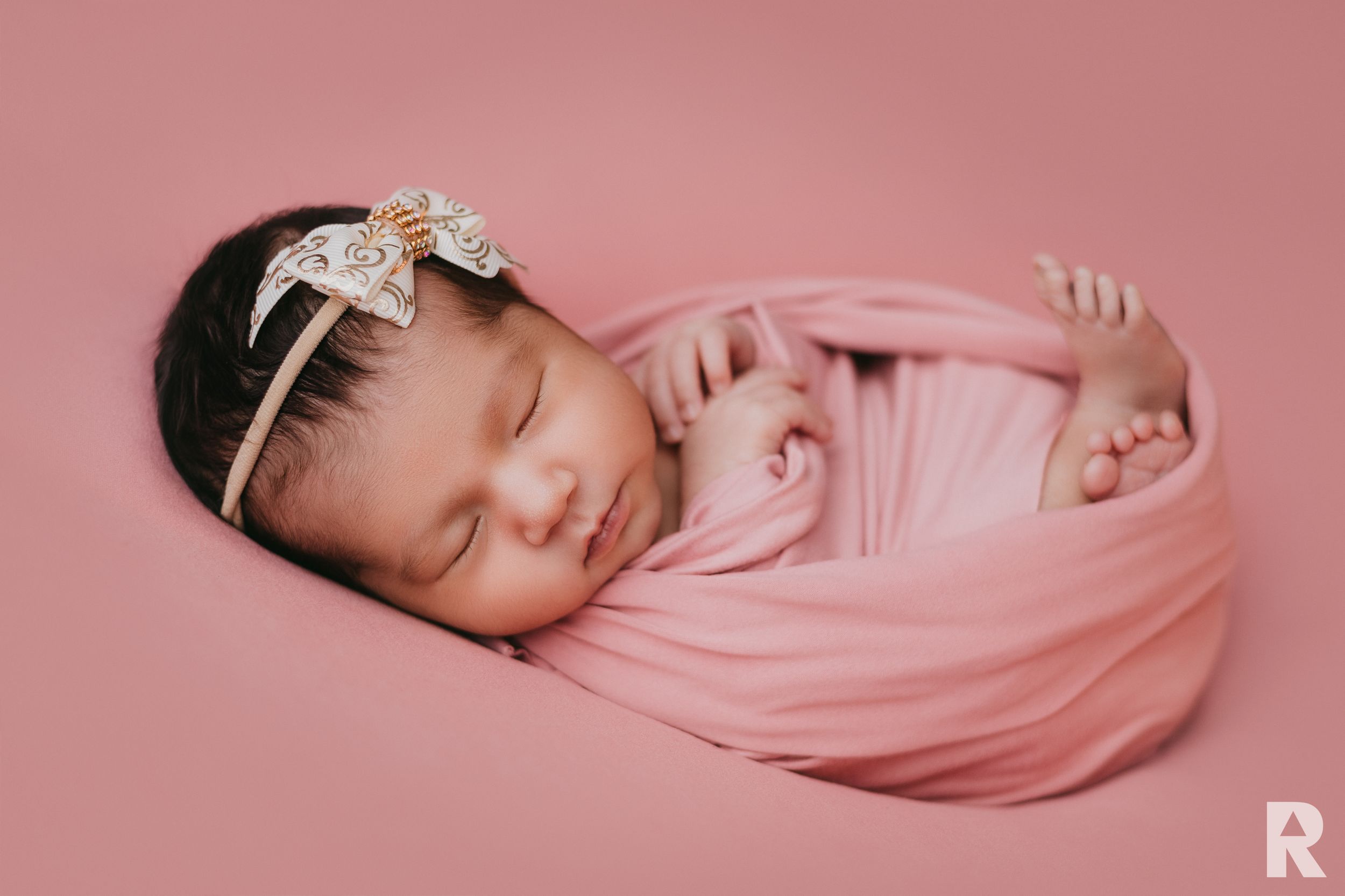 Sleeping newborn wrapped in soft pink swaddle on pink background with delicate headband.