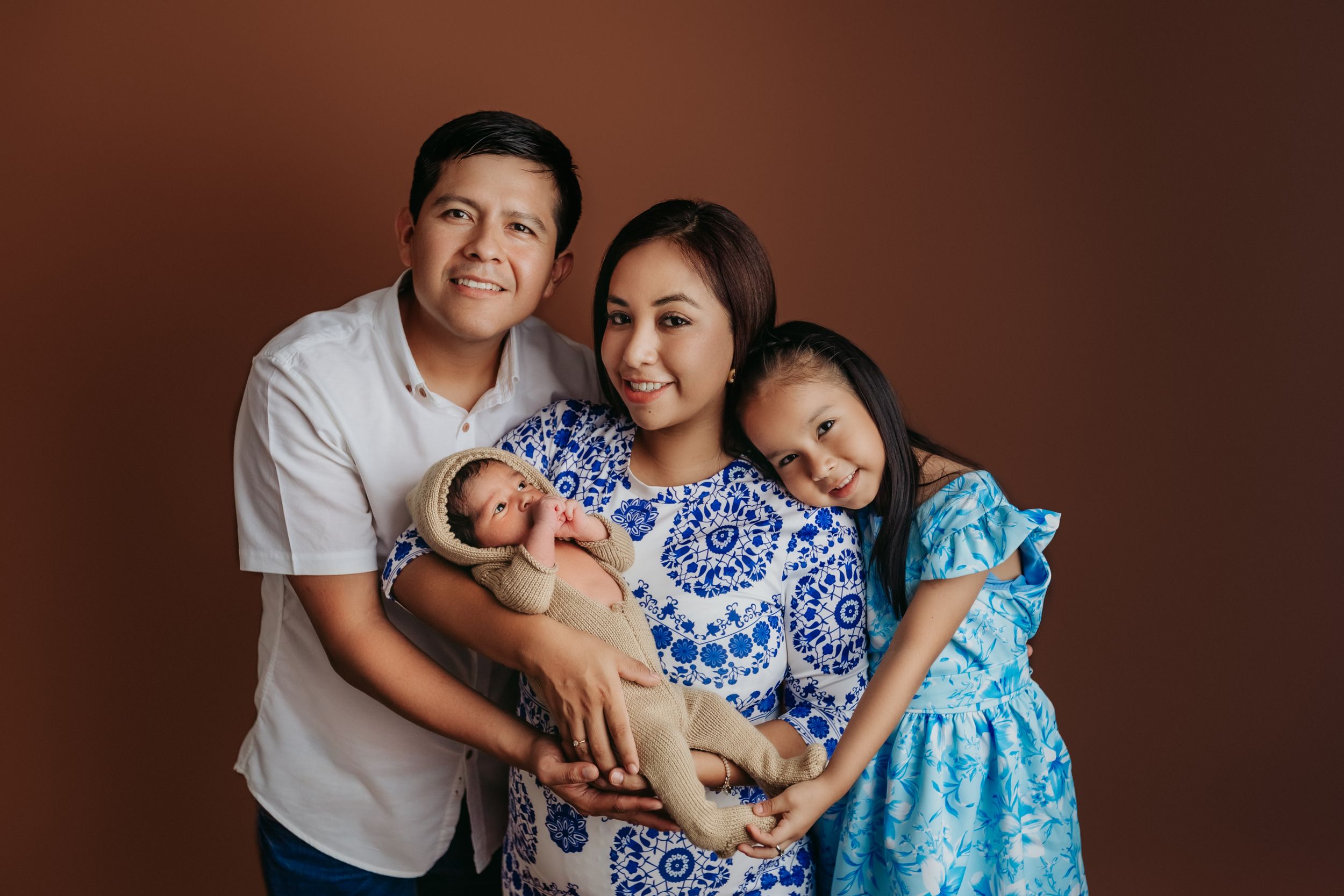 Beautiful family portrait against brown backdrop shows loving embrace with newborn.