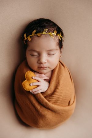 Sleeping newborn with gold leaf crown wrapped in caramel colored blanket holding plush toy.