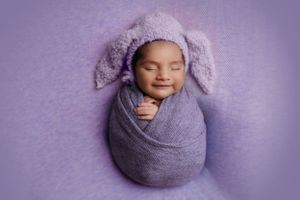 Newborn wearing purple bunny ear hat wrapped in cozy blanket against purple background.