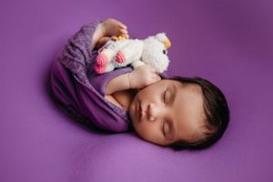 Sleeping newborn wrapped in purple swaddle holding white plush toy on purple background.