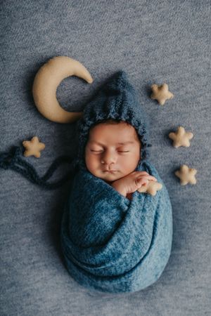 Sleeping newborn in blue wrap with knit hat surrounded by decorative moon and stars on gray backdrop.