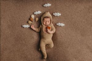 Adorable newborn in brown knit bear outfit lying on brown backdrop with decorative white paper clouds.