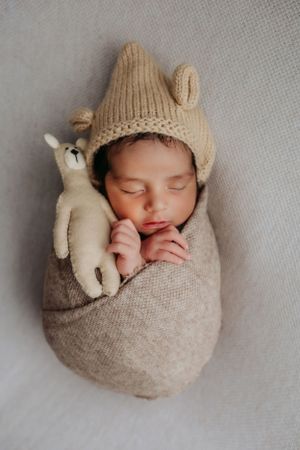 Sleeping newborn in beige knit bunny hat and wrap snuggling with plush toy on white background.