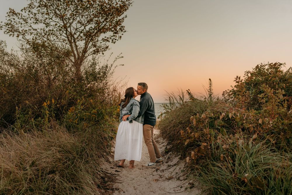 Couple stands together on a coastal trail during sunset with natural foliage surrounding them.