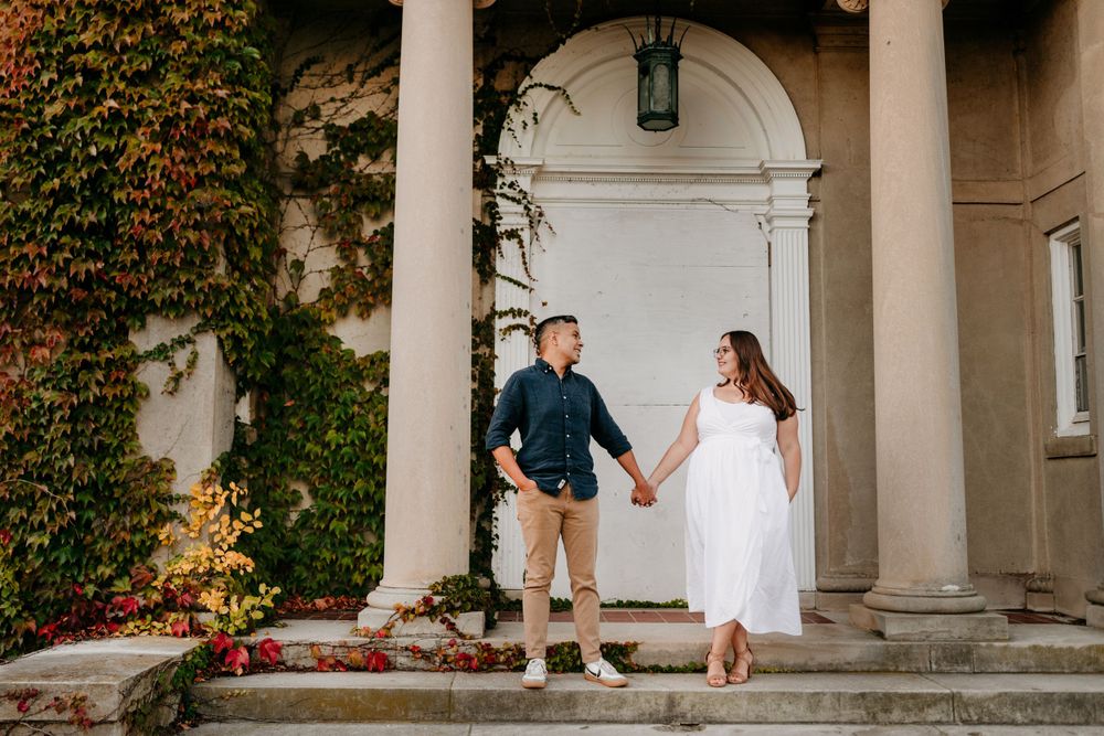 A couple poses on a columned porch with ivy-covered walls and classic architectural details.