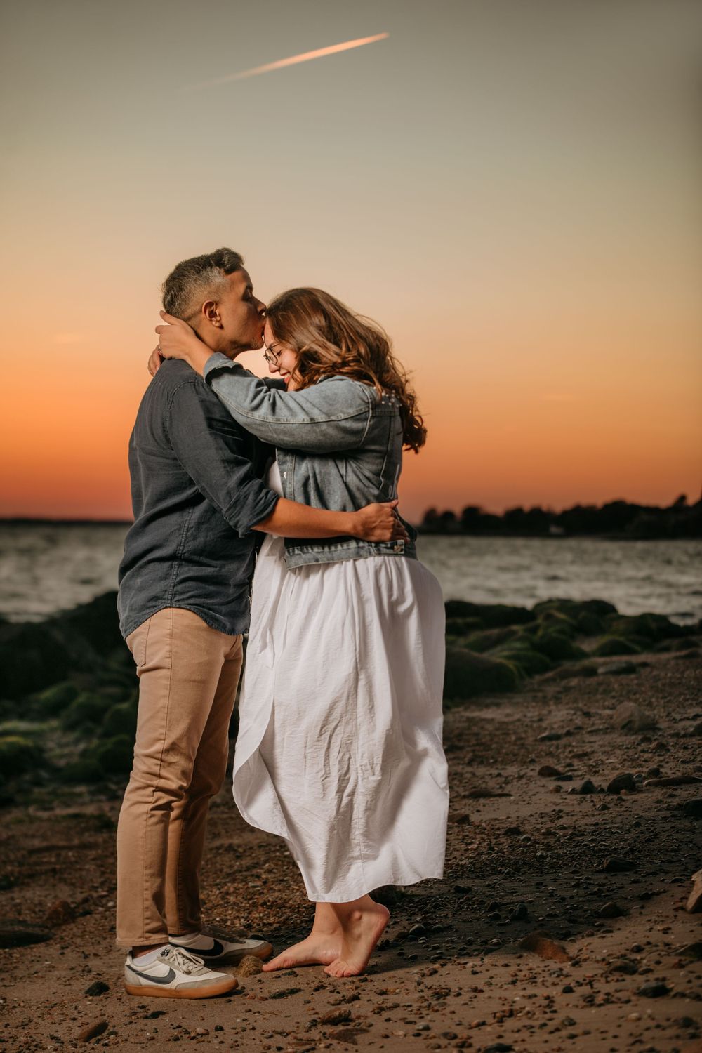 Couple embraces on a beach during a dramatic orange sunset with waves crashing behind them.