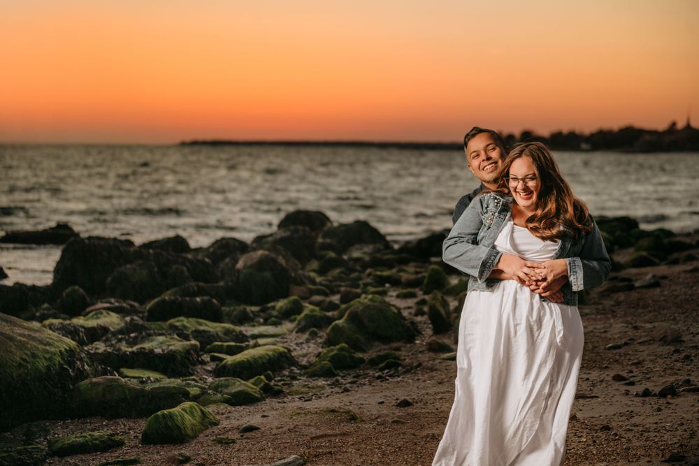 Couple shares intimate embrace on rocky beach shore during vibrant orange sunset.