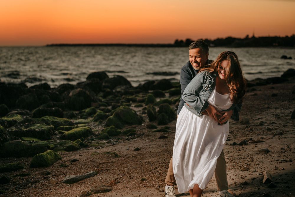 Romantic couple embraces on rocky beach during vibrant orange sunset along coastline.