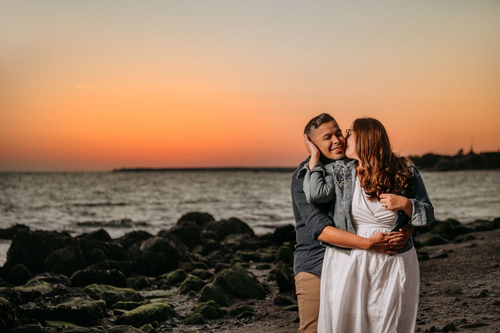 Intimate moment shared on beach with dramatic orange sky and rocky shoreline.