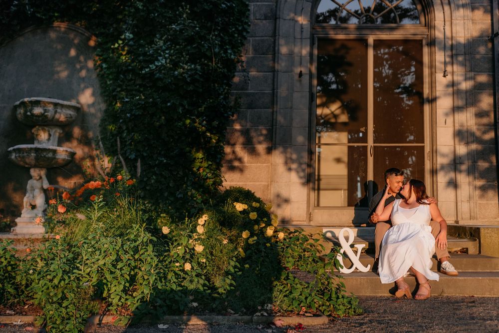 A couple shares a playful moment in front of a sunlit building with lush garden scenery.