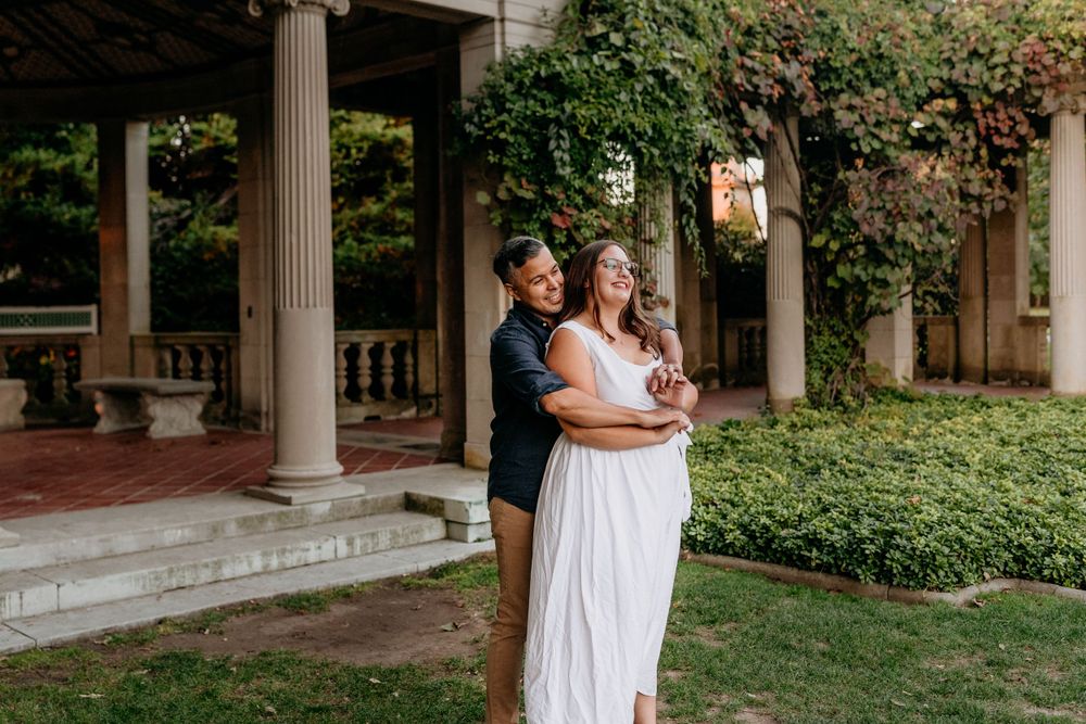 A couple embraces under an elegant columned portico surrounded by lush greenery and climbing vines.