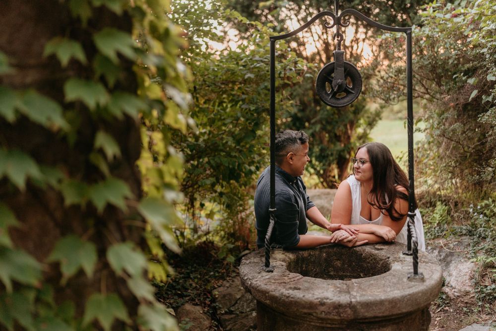Couple shares an intimate moment while sitting on a stone well surrounded by ivy and garden foliage.