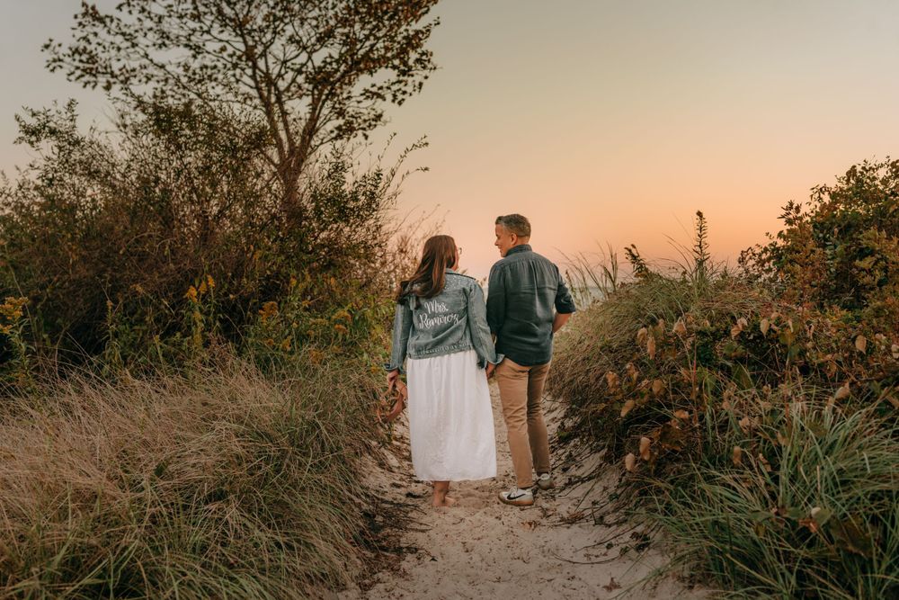 Couple walks hand in hand on a sandy path during golden hour surrounded by tall grass.