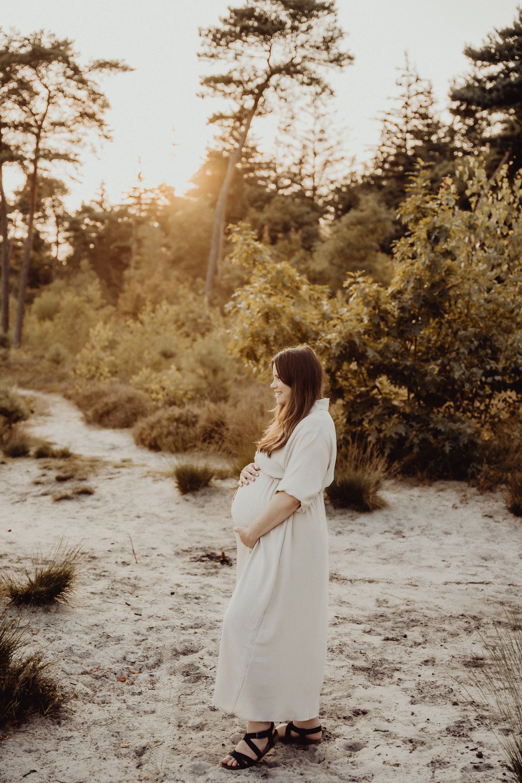 Peaceful maternity photoshoot in a sunlit forest with flowing white dress.
