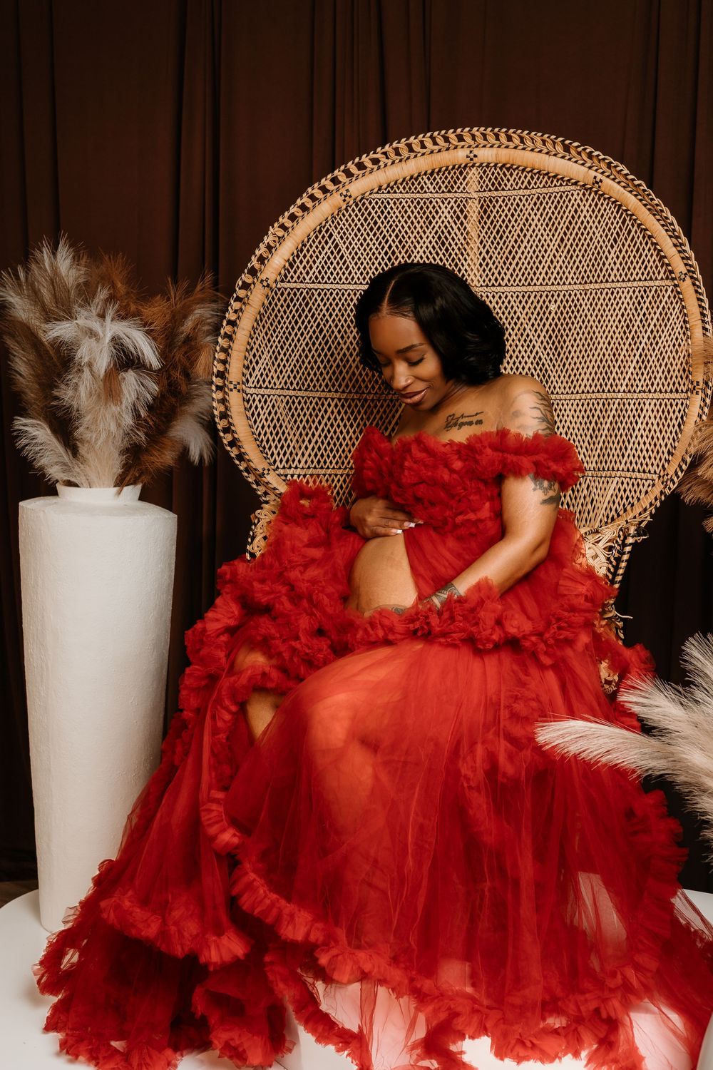 A person in a flowing red tulle gown poses dramatically on a wicker peacock chair against a dark background.