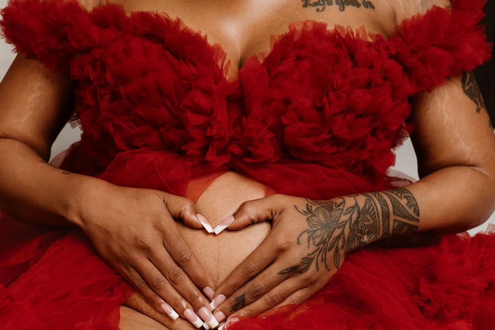 A person in a red ruffled dress holds their hands over their stomach, with one arm displaying a floral tattoo.