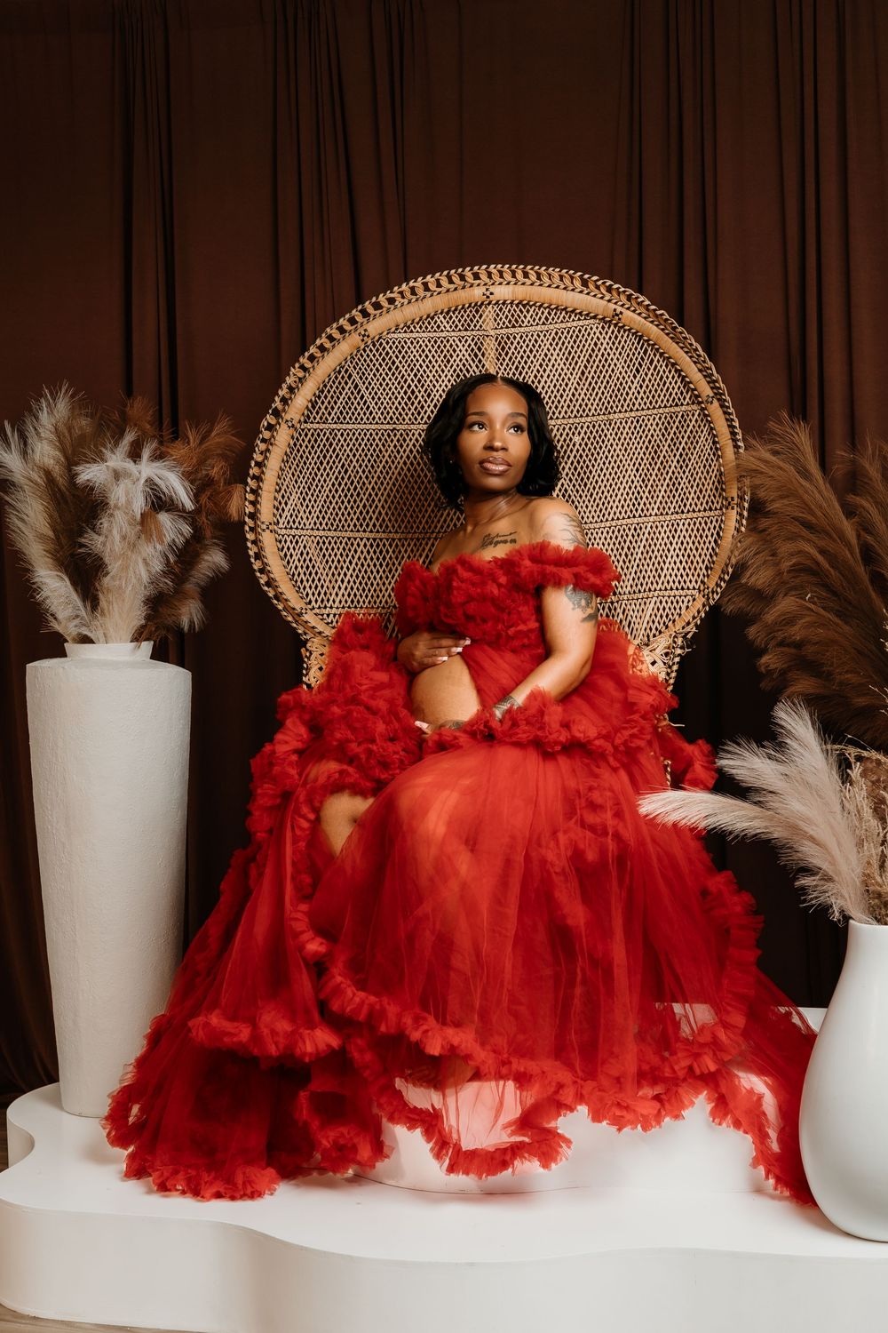 A glamorous portrait session featuring a red tulle gown, wicker peacock chair, and white pampas grass decor.