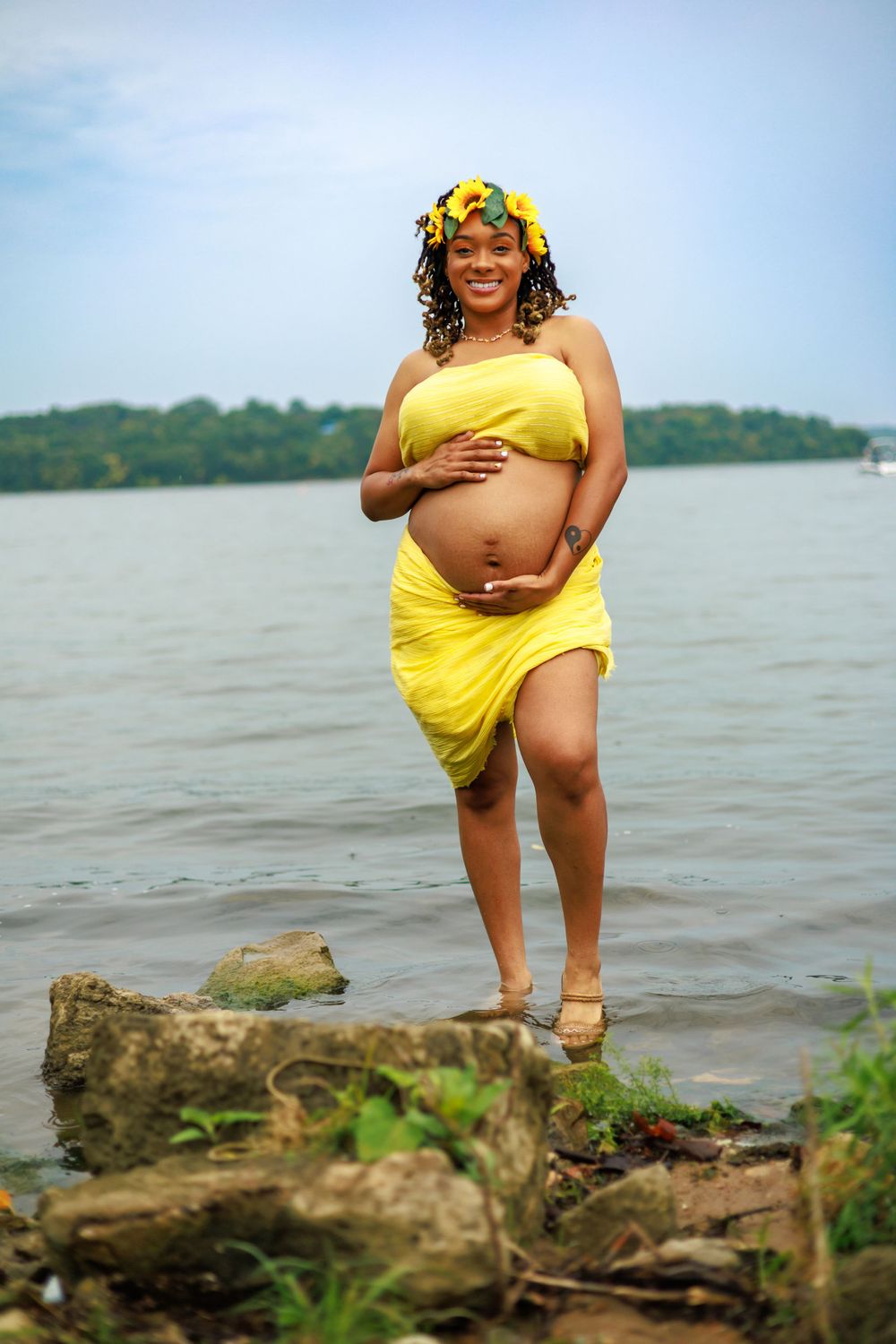 Pregnant woman in yellow dress poses by waterfront with scenic background.