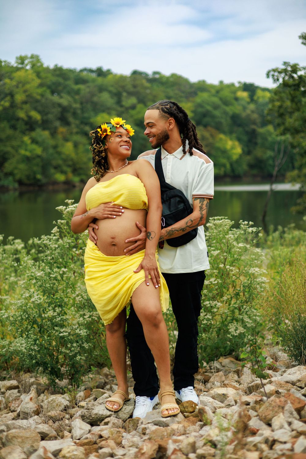 Expecting couple shares intimate moment by lake with sunflowers in hair.