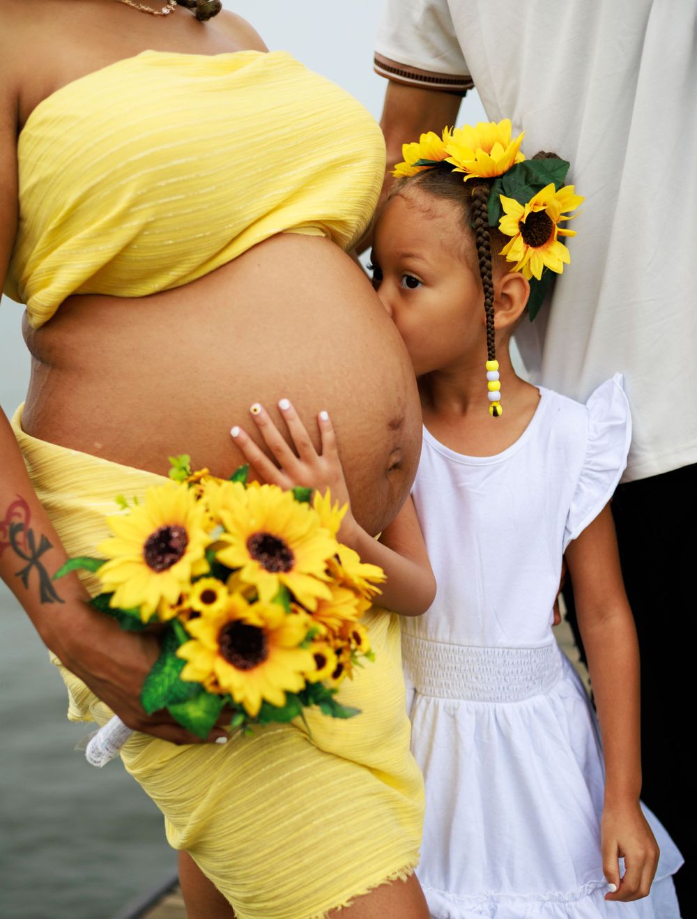 Maternity photo featuring yellow dress and white outfit with sunflower decorations against dark background.