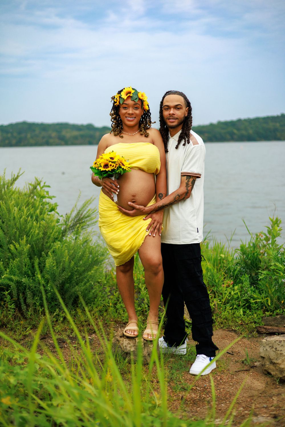 Maternity photo with yellow dress and sunflower crown against lakeside backdrop.
