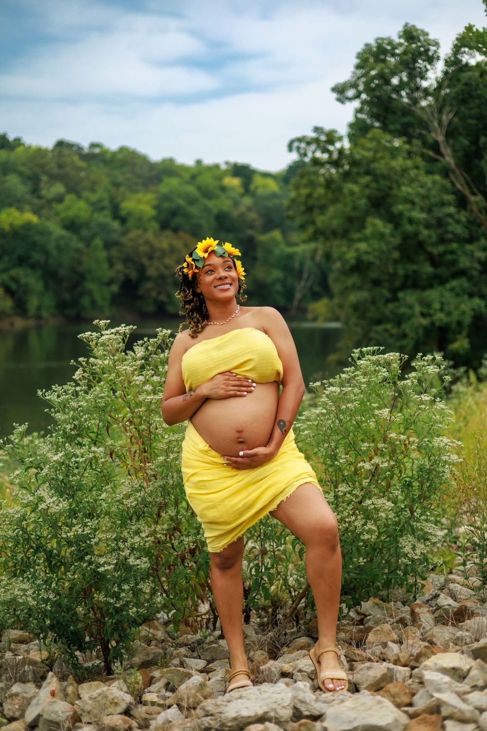 An expectant mother in yellow poses confidently by a lake, wearing a sunflower crown among wildflowers.